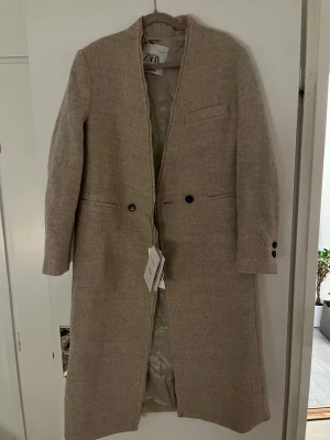 Beige kappa från Zara i ullmix - Stilren beige kappa från Zara i ullmix från Manteco. Lång modell med rak passform, knappar framtill och diskreta fickor. klassisk look som passar perfekt till höst och vinter. Snygga knappdetaljer vid ärmslut.