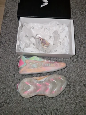 AE 1 low basket skor - Säljer ett par riktigt snygga Adidas AE 1 basket skori pastellfärger med inslag av rosa, blått och gult. Skorna har meshdetaljer på sidorna, vit snörning och en genomskinlig sula med rosa detaljer. Adidas-logga i vitt på hälen och cool futuristisk design. Vanliga priset går för 1300 men säljer fär billigare