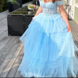 Ljusblå tyllklänning med volanger - Drömmig långklänning i ljusblå tyll med flera lager volanger och offshoulder-liv. Överdelen har spetsdetaljer och markerad midja, vilket ger en sagolik känsla. Perfekt för bal. Har använts två vällar och är lite sliten underkjolen  men är inget som syns. Som ni ser på en av bilderna så är en av ”padsen” sönder vid bysten men det är inte heller något som syns.
