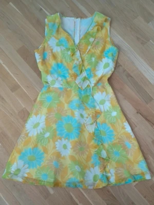 Blommig klänning - Söt kortklänning med blommigt mönster i gult, blått, grönt och vitt. Klänningen har omlottdetalj framtill med volang längs kanten och är ärmlös.