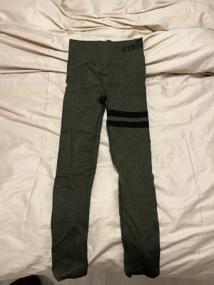 Mörkgröna leggings med svarta ränder - Snygga mörkgröna leggings med två svarta ränder på ena benet och bred resår i midjan. Perfekta för träning eller chill, med stretchigt material som sitter skönt. Diskret text upptill vid midjan.