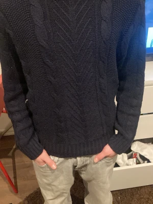 Mörkblå stickad tröja från Leftiesman - Mörkblå stickad tröja från Leftiesman i storlek S. Tröjan har ett snyggt kabelstickat mönster framtill och på ärmarna, samt ribbade muddar vid ärmslut och nederkant. Perfekt för kyliga dagar och enkel att matcha med jeans.