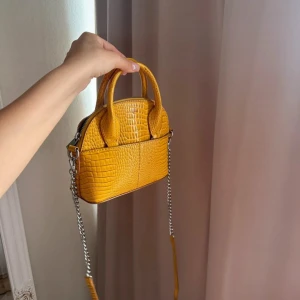 Gul väska med kedja - Säljer en gul väska i croco-mönstrat skinnimitation med silverfärgad kedja och dragkedja. Väskan har både handtag och axelrem.