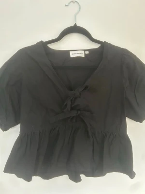 Svart blus med knyt från Carin Wester - Supersnygg svart blus från Carin Wester i storlek 36. Modellen har korta puffärmar, v-ringning och två dekorativa knyt framtill. Blusen har en volangkant nedtill som ger en luftig och trendig look. Perfekt att styla med jeans eller kjol.