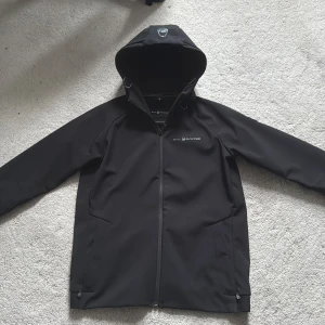 Svart vindjacka från Sail Racing - Säljer en svart vindjacka från Sail Racing med huva och dragkedja framtill. Jackan har en stilren design med logga på bröstet och smarta detaljer som dragsko i huvan. Perfekt för dig som vill ha en snygg och funktionell jacka till vardags.