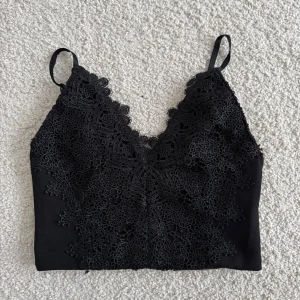 Svart spetslinnetopp från BikBok - Säljer en svart linnetopp från BikBok i storlek XS. Toppen har tunna axelband, v-ringning och är dekorerad med detaljerad spets framtill. Materialet känns mjukt och stretchigt, perfekt för att styla med höga jeans eller kjol.