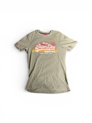 Grön Superdry t-shirt med tryck - Säljer en grön t-shirt från Superdry med coolt retrotryck i vitt, rött och gult på bröstet. Klassisk passform och rund hals. Perfekt för dig som gillar streetwear och vill ha något bekvämt och stilrent. Materialet är mjuk bomull.