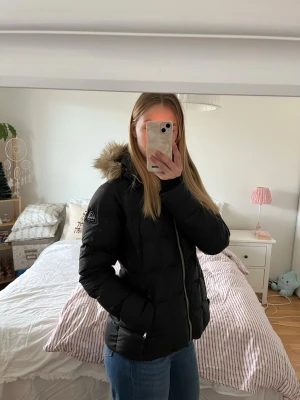 Svart dunjacka med fuskpäls från McKinley - Säljer en svart dunjacka från McKinley med huva och fuskpäls. Jackan har quiltad design, dragkedja framtill och två fickor med dragkedja. Perfekt för kalla dagar och har en klassisk look som funkar till det mesta.