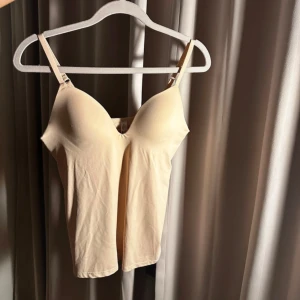 Seamless topp - Säljer en beige väst med smala, justerbara axelband och vadderade kupor. Västen har en enkel, slät design och är tillverkad i ett mjukt syntetmaterial som känns skönt mot huden. Perfekt att styla under en skjorta eller som den är till jeans.