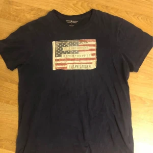 Mörkblå t-shirt från Ralph Lauren - Säljer en mörkblå t-shirt från Ralph Lauren med ett stort tryck av en amerikansk flagga och texten 'Denim & Supply' på framsidan. T-shirten är tillverkad i 100% bomull och har en klassisk passform.