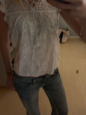 Vit broderad blus med knyt - Säljer en söt vit blus i bomull med broderade detaljer och hålmönster. Blusen har korta ärmar, volangkant och knytband med tofsar vid halsen. Perfekt till sommaren och ger en romantisk vibe till din outfit. Använd 1 gång 💞💞