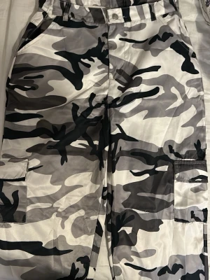 Cargopants i grått camomönster - Säljer ett par cargopants med grå, svart och vit camouflage. Byxorna har stora fickor på sidorna och klassisk cargomodell. Materialet känns lätt och lite glansigt, perfekt för dig som vill sticka ut med streetwear vibe.