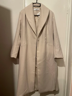 Beige kappa från Ellos - Säljer en stilren beige kappa från Ellos med bred krage och knappar framtill. Långärmad modell med fickor på sidorna och rak passform. Perfekt för dig som vill ha en klassisk och enkel look under kyligare dagar.