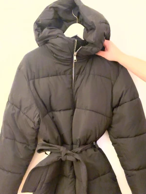 Svart pufferjacka med bälte från Nelly - Säljer en svart pufferjacka från Nelly med stor huva och bälte i midjan för snygg siluett. Jackan har dragkedja framtill och quiltad design som ger en riktigt mysig känsla. Perfekt för kalla dagar när du vill vara både varm och stilren. !!ANVÄND MAX 3 GÅNGER!! Storlek XS men ganska stor i storlek