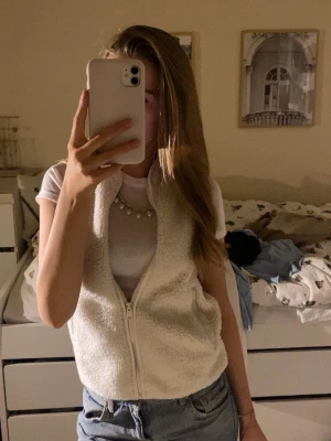 Vit teddyväst med dragkedja - Säljer en mysig vit teddyväst med dragkedja framtill. Västen är ärmlös och har en fluffig, mjuk yta som ger en chill vibe. Perfekt att slänga över en t-shirt eller hoodie för en avslappnad look. Passar till jeans eller kjol för en trendig stil.