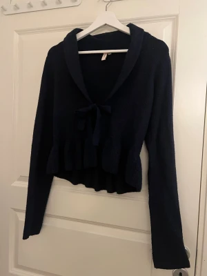 Mörkblå stickad kofta från Nelly - Säljer en mörkblå stickad kofta från Nelly i storlek XS. Koftan har lång ärm, volangkant nedtill och en snygg knytning framtill vid halsen. Perfekt för dig som gillar en feminin och trendig look med lite extra detaljer.