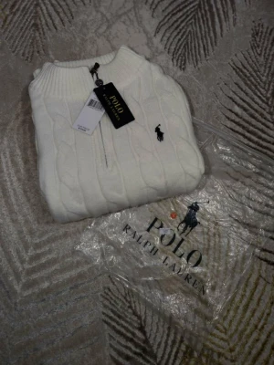 Vit kabelstickad tröja Polo Ralph Lauren - Vit kabelstickad tröja från Polo Ralph Lauren med klassisk svart logga på bröstet. Tröjan har ribbade muddar, hög krage och dragkedja upptill. Perfekt för dig som gillar stilrena och tidlösa plagg.