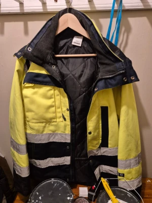 Gul och svart vinterjacka Fristads XL - Säljer en gul och svart vinterjacka från Fristads i storlek XL. Jackan har reflexdetaljer, flera fickor, dragkedja och tryckknappar. Insidan är quiltad och jackan har hög krage och justerbar huva. Perfekt för kalla dagar och synlighet utomhus.
