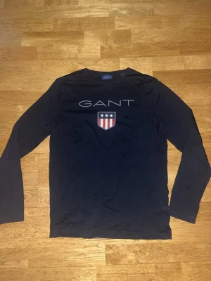 Svart långärmad tröja från GANT - Säljer en svart långärmad tröja från GANT med stort tryck på bröstet och ett sköldmärke med stjärnor och ränder. Tröjan har rund hals och är gjord i mjuk bomull, perfekt för en clean och stilren look.