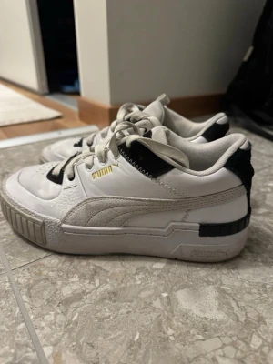 Vita sneakers från Puma med svarta detaljer - Säljer ett par snygga vita Puma sneakers med svarta detaljer och guldfärgad Puma-logga på sidan. Skorna har klassisk snörning, rund tå och en chunky sula. Materialet är en mix av skinn och mocka vilket ger en schysst kontrast. 