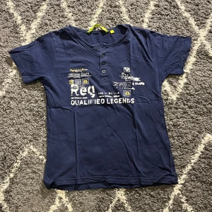 Mörkblå t-shirt med tryck och knappar - Snygg mörkblå t-shirt i 100% bomull med coolt tryck framtill och texten 'Qualified Legends'. Storlek är 152. T-shirten har tre knappar vid halsen för en extra detalj och korta ärmar. Perfekt för en avslappnad och sportig look.