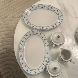 Porslinsservis med blå rosdekor - Elegant porslinsservis med två ovala fat, två sockerskålar med lock och en gräddkanna. Vit bas med blå rosdekor och blå kant. Alla delar ser ut att vara i mycket gott skick utan synligt slitage eller sprickor. Perfekt för servering och dukning med klassisk stil.