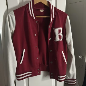 Röd och vit varsityjacka med B - Snygg varsityjacka i rött och vitt med stora B-applikationer på bröstet och broderad text 'Varsity Team 28' på ärmen. Jackan har ribbade muddar med vita ränder, tryckknappar och två fickor framtill. Materialet är troligtvis bomull och polyester. Kommer inte till användning ‼️ använt 1 gång för många år sedan ☺️ Skit snygg ☺️‼️