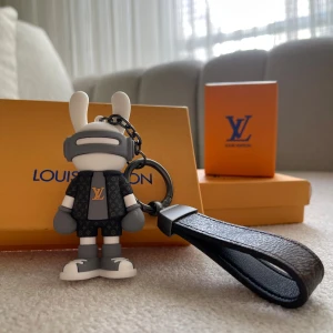 Vuitton nyckelring med figur - Exklusiv Vuitton nyckelring med en cool figur i grått och vitt, klädd i monogrammönstrad outfit och sneakers. Nyckelringen har en brun läderrem med klassiskt mönster och metallfäste. Perfekt accessoar för dig som gillar designer och streetstyle.