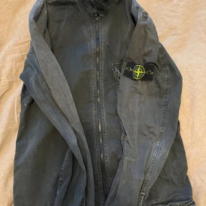 Svart overshirt från Stone Island - Svart overshirt från Stone Island med klassisk logopatch på ärmen. Jackan har dragkedja framtill och en stilren krage. Materialet är mjukt och lätt, perfekt för lager-på-lager. Snygga detaljer och ikonisk design som ger en cool streetkänsla.