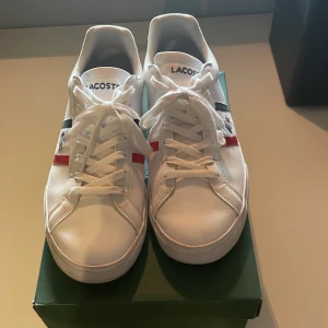 Vita Lacoste sneakers med röd och blå detalj - Snygga vita sneakers från Lacoste med klassisk krokodillogo på sidan. Skorna har snörning och coola röd-blå ränder längs sidan för extra stilpoäng. Tillverkade i skinn med platt sula och rund tå. Perfekta för dig som gillar sportig och fräsch look. Storlek 43. Nypris 1200kr mitt pris 700