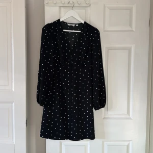 Svart prickig kortklänning från Kappahl - Svart kortklänning från Kappahl med vita prickar, v-ringad hals och knappar framtill. Klänningen har långa ärmar med lätt puff och är tillverkad i 100% återvunnen polyester. Perfekt för dig som gillar klassiska mönster med en modern twist.