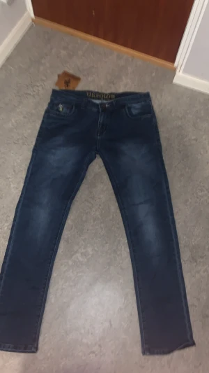 Mörkblå jeans från U.S. Polo Assn - Snygga mörkblå jeans från U.S. Polo Assn med broderad logga i midjan och klassiska fem fickor. Jeansen har smal passform och är tillverkade i stretchigt denimtyg för extra komfort. Perfekta för en trendig och avslappnad look.