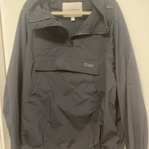 Svart vindjacka från Carin Wester - Svart vindjacka från Carin Wester i stilren design med halv dragkedja och stor ficka framtill med CW-logga. Jackan har justerbar huva med snörning och är långärmad. Perfekt för dig som gillar minimalistisk och funktionell stil.