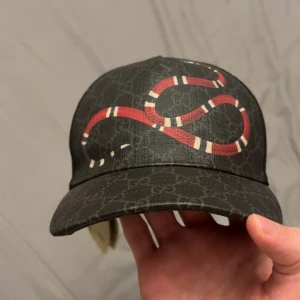 Gucci keps med röd orm och GG-mönster - Snygg svart keps från Gucci med klassiskt GG-mönster och en fet röd och vit orm på framsidan. Baksidan har mesh och justerbart band med röd och blå detalj. Perfekt för dig som vill sticka ut med designer vibes.