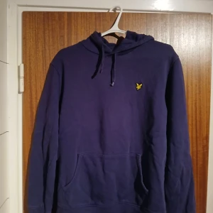 Mörkblå hoodie från Lyle & Scott - Säljer en mörkblå hoodie från Lyle & Scott med klassisk gul logga på bröstet. Tröjan har huva med snörning och en stor magficka. Perfekt för dig som gillar en enkel och stilren look.