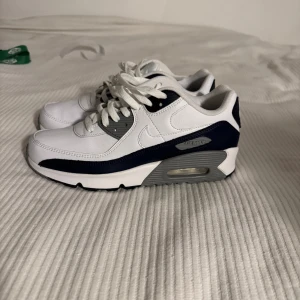 Nike Air Max 90 vit och marinblå - Nike Air Max 90 sneakers i vit och marinblå med grå detaljer. Klassisk design med synlig Air-enhet i sulan, rund tå och snörning. Ovandel i syntet och skinn, platt sula och ikonisk Swoosh på sidan. Perfekt för dig som gillar stilrena och sportiga sneakers.