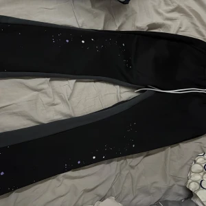 Svarta SUM WON mjukisbyxor med stänk - Svarta mjukisbyxor från SUM WON med grå sidopaneler och vita samt lila färgstänk längs benen. Byxorna har snörning i midjan och är tillverkade i mjukt syntetmaterial, perfekt för chill eller streetwear. Snygg och unik design med konstnärlig touch.