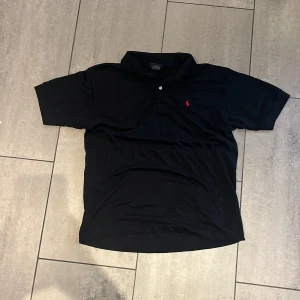 Svart Polo Sport pikétröja L - Svart pikétröja från Polo Sport i storlek L, tillverkad i 100% bomull. Klassisk krage, korta ärmar och en röd broderad logga på bröstet. Perfekt för dig som gillar stilrena och sportiga plagg med en enkel men snygg look.