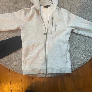 Grå hoodie från Sail Racing med huva - Snygg ljusgrå hoodie från Sail Racing med huva och hel dragkedja framtill. Tröjan har en bröstficka med dragkedja och diskret logga på vänster sida. Perfekt för chill dagar och har en skön passform med långa ärmar. 170 cm! Passar mig perfekt då jag är 175 cm lång och väger 55 kg! Skriv vid frågor!