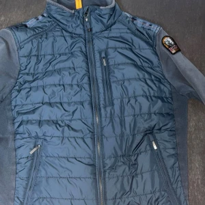 Blå pufferjacka från Parajumpers - Säljer en blå pufferjacka från Parajumpers i storlek L. Jackan har quiltad framsida, fleecepartier på ärmar och sidor, samt dragkedjor på fickor och bröst. Märkespatch på axeln och ståkrage med orange detalj. Perfekt för dig som gillar sportig stil.