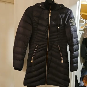 Svart lång dunjacka från bubbelroom  - Säljer en svart lång dunjacka från bubbelroom med huva och snygga guldfärgade dragkedjor. Jackan har quiltad design, två fickor med dragkedja och en patch med logga på ärmen. Perfekt för kalla dagar och har en modern, stilren look.