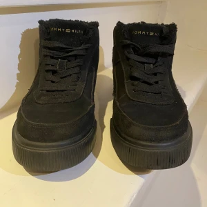 Svarta sneakers Tommy Hilfiger mocka - Svarta sneakers från Tommy Hilfiger i mocka med högre skaft och platt sula. Ca 1000 kr, använda i en säsong utan märken, inga skador. Skorna har snörning och diskret logga på sidan och bak. Insidan är fodrad med mjukt material och yttersulan är räfflad för extra grepp. Perfekt för dig som gillar stilrena och bekväma skor.