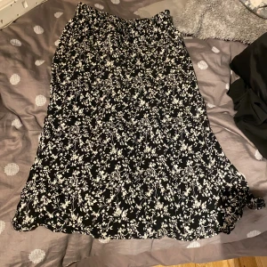 Svart blommig långkjol från 157 - Svart långkjol med vitt blommigt mönster från 157 i storlek XS. Kjolen har en flowy passform och är gjord i ett lätt och mjukt material, perfekt för dig som gillar en chill och feminin stil. Elastisk midja för extra komfort.