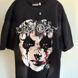 Svart Hellstar t-shirt med tryck - Svart t-shirt från Hellstar med stor grafisk print av ett ansikte med taggtråd och färgstänk på framsidan. Baksidan har Hellstar-loggor i rosa, gult och vitt. Oversized passform och klassisk rund hals. Perfekt statement-plagg för dig som gillar streetwear.