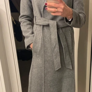Grå kappa med bälte från H&M - Snygg grå kappa från H&M i storlek 32. Kappan har ett stilrent bälte i midjan, långa ärmar och praktiska fickor. Materialet är en mix av polyester, ull och viskos som ger en mjuk och klassisk känsla. Perfekt för dig som gillar minimalistisk och clean stil.