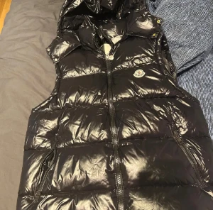 Svart dunväst från Moncler - Svart glansig dunväst från Moncler med huva och dragkedja framtill. Västen har två sidofickor med dragkedja och ikonisk Moncler-logga på bröstet. Insidan har ett serietidningsmotiv och märkestaggar. Perfekt för att addera en cool vibe till din outfit.