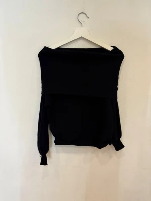 Svart offshoulder stickad tröja - Svart stickad tröja med offshoulder-design och långa ärmar. Tröjan har en bred krage som vilar över axlarna och ger en cool, avslappnad vibe. Perfekt för dig som gillar stilrena och enkla plagg med en twist.