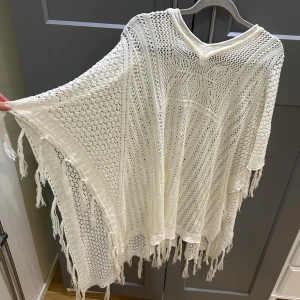 Vit stickad poncho från MinMilo - Säljer en vit stickad bolerotopp/ poncho från MinMilo i storlek L/XL. Toppen har ett luftigt hålmönster, v-ringad hals och fransar längs kanterna för en boho-vibe. 