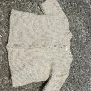 Beige stickad kofta med knappar kortärmad - Mysig beige stickad kofta med rund hals och långa ärmar. Koftan har ribbade muddar och fem knappar framtill. Perfekt för lager-på-lager och chill dagar. Enkel och stilren design som funkar till det mesta. En liten fläck där bak men inget man tänker på