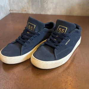 Mörkblå sneakers från Tretorn - Snygga mörkblå sneakers från Tretorn med gulddetaljer och vit platt sula. Skorna har snörning och är tillverkade i mocka med låg profil. Tretorn-logga i guld på sidan och hälen, samt '55' på plösen. Perfekta för dig som gillar stilrena och klassiska sneakers.